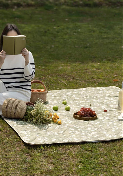 Person reading a book outdoors on a picnic blanket with fruits and flowers.